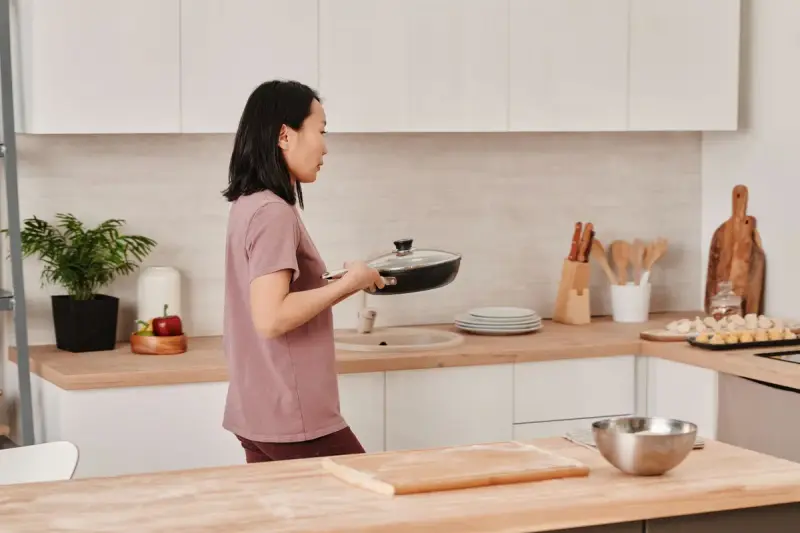 Woman carrying a lidded frying pan across a bright modern kitchen with wooden counters