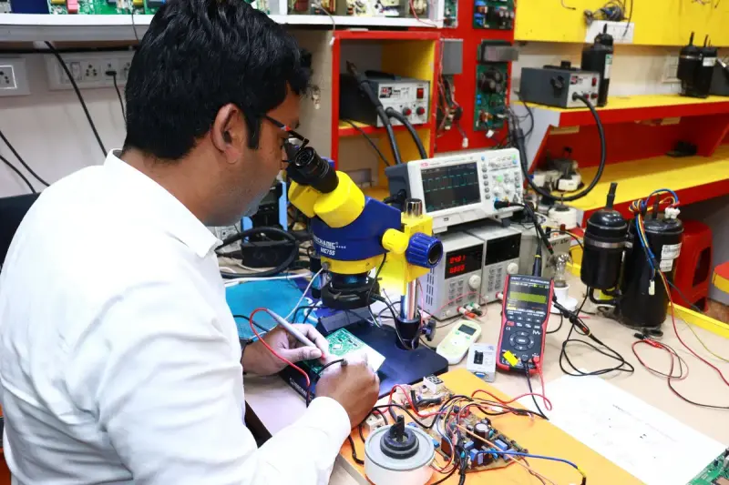 A technician works on a circuit board under a microscope in an electronics lab