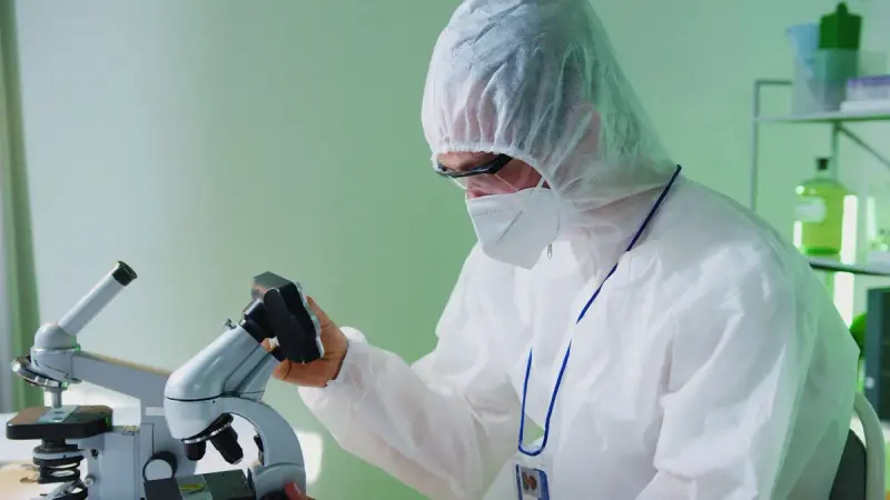 A scientist in protective gear looks into a microscope in a laboratory