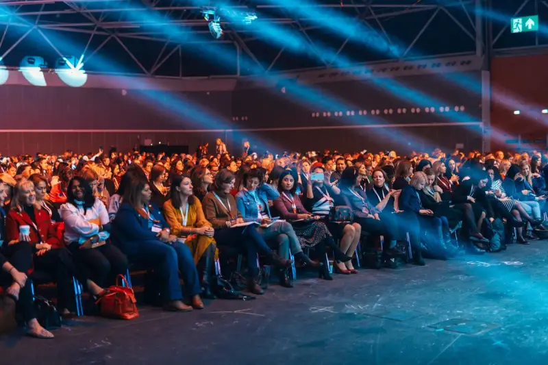 A large seated audience in an auditorium under colored stage lighting
