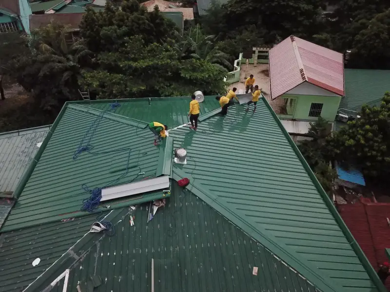 Aerial view of a crew installing green metal roof panels on a house among trees