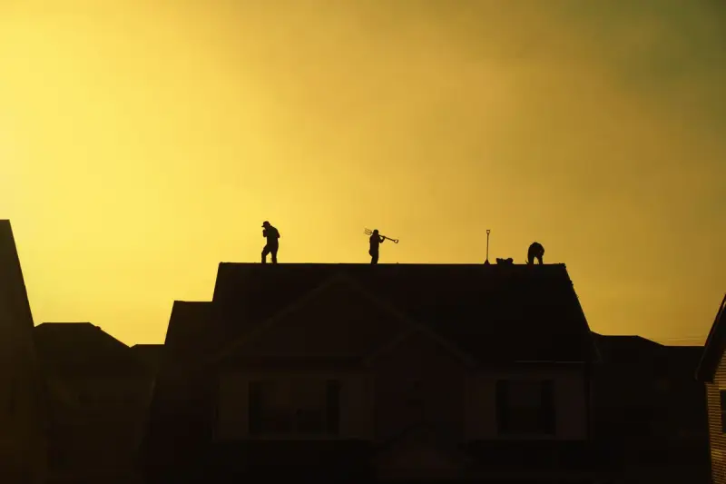 Three roofers silhouetted on top of a suburban house against a golden sunset sky
