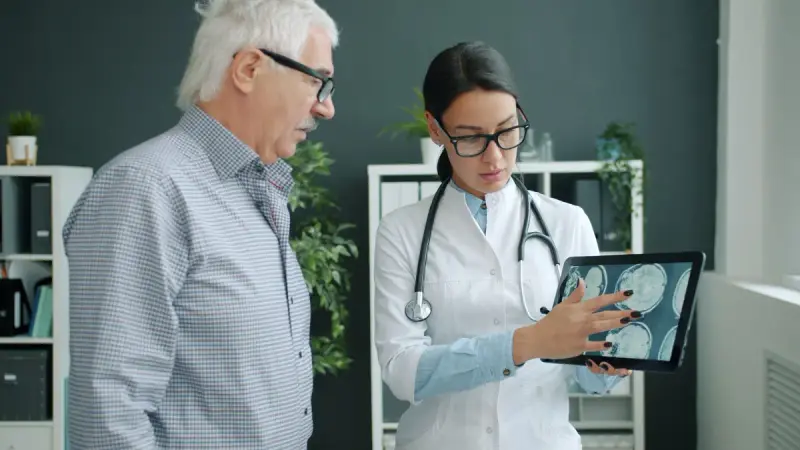 Woman doctor in a white coat showing brain scans on a tablet to an older man