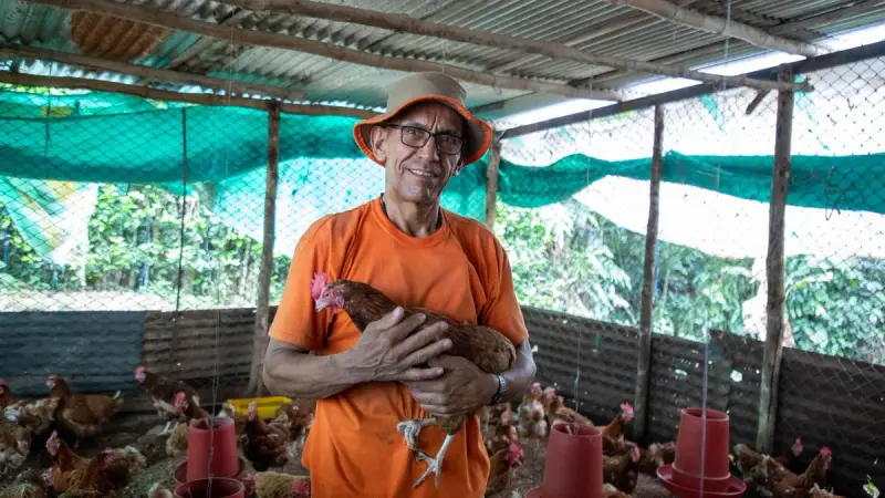 Smiling poultry farmer in an orange shirt holding a brown hen inside a shaded poultry house with feeders
