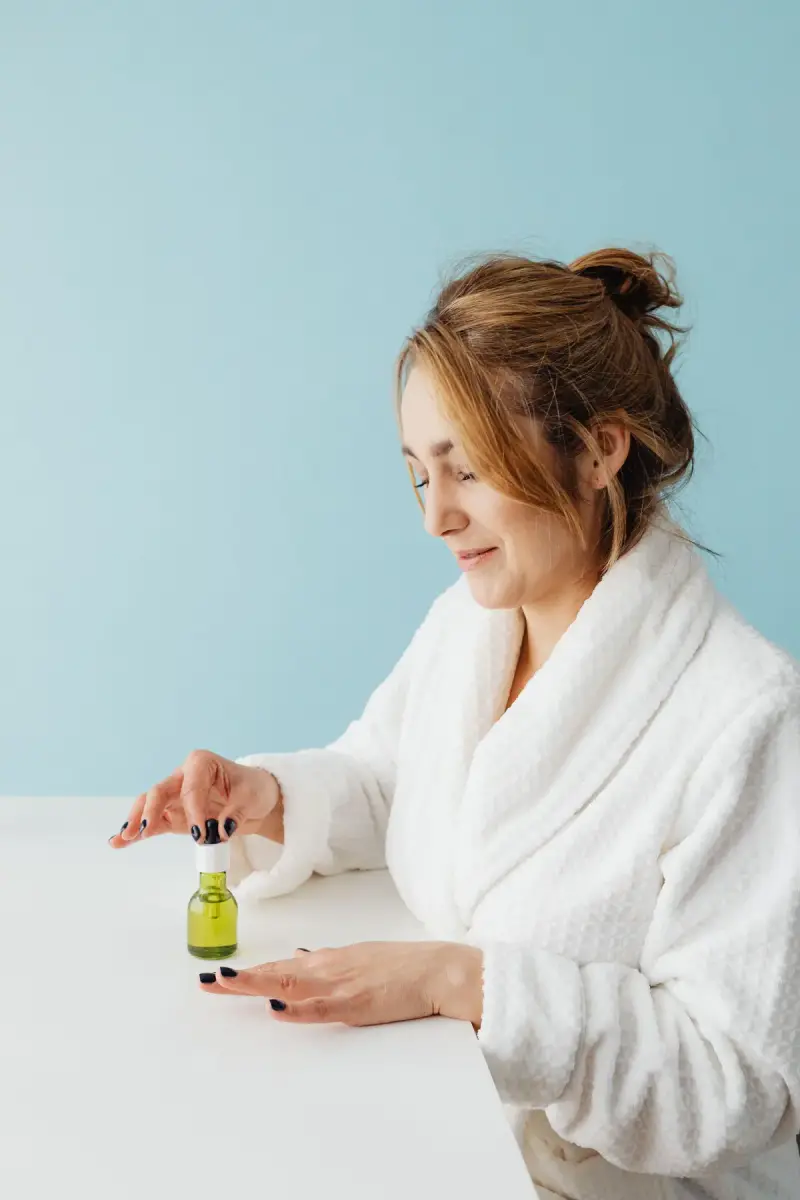 Woman in a white bathrobe opening a green serum bottle at a table