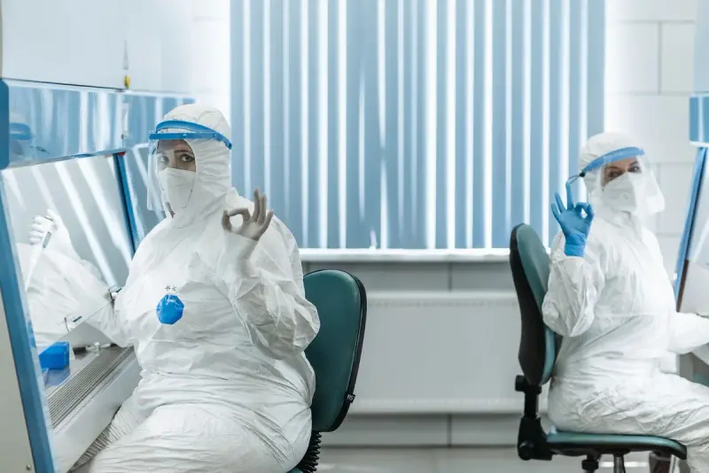 Two operators in full white gowns, masks and face shields at biosafety cabinets in a bright lab