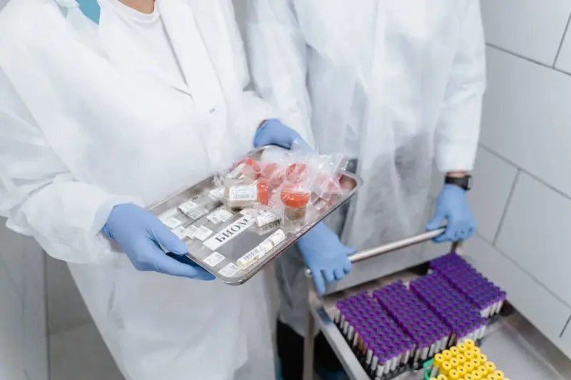 Two lab workers in gowns and gloves carrying a steel tray of samples and a rack of tubes