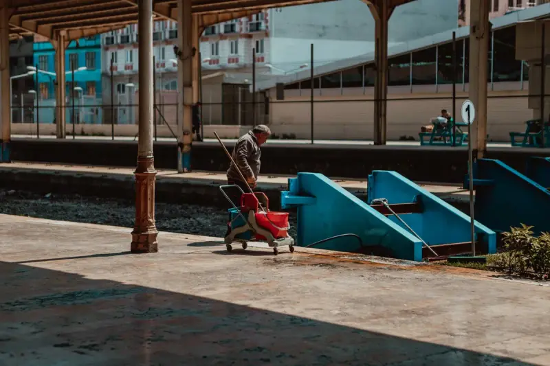 Worker pushing a cleaning cart along a sunlit railway station platform