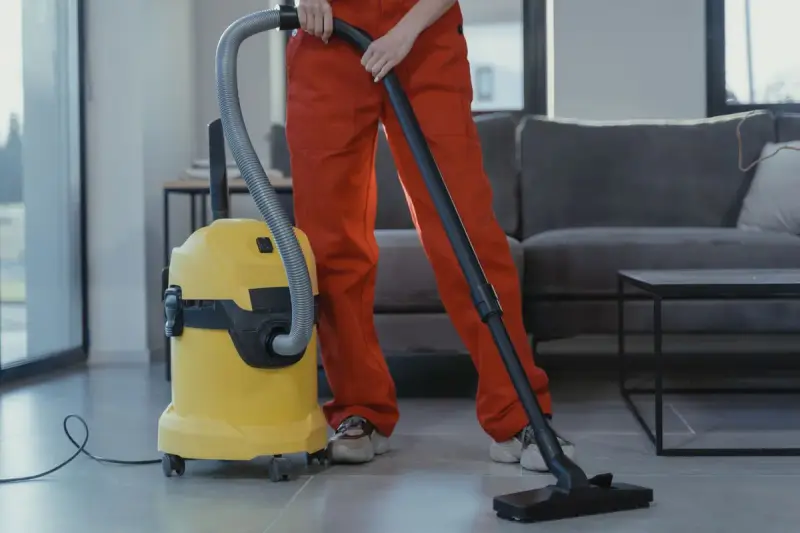 Cleaner in red coveralls vacuuming a concrete floor with a yellow wet-dry vacuum