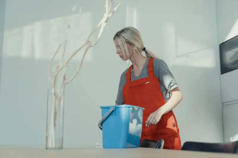 Cleaner in a red apron lifting a blue bucket in a bright white room