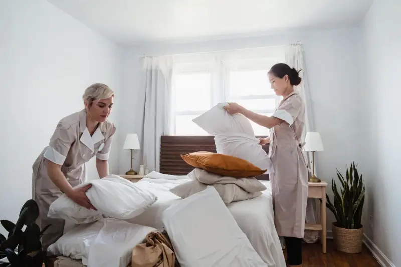 Two hotel housekeepers in uniform arranging pillows and fresh bedding on a hotel bed