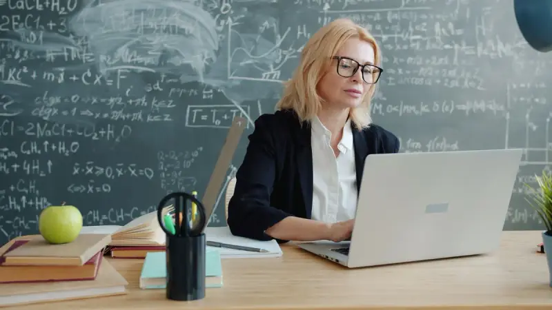 Educator working on a laptop in a classroom with equations on the chalkboard