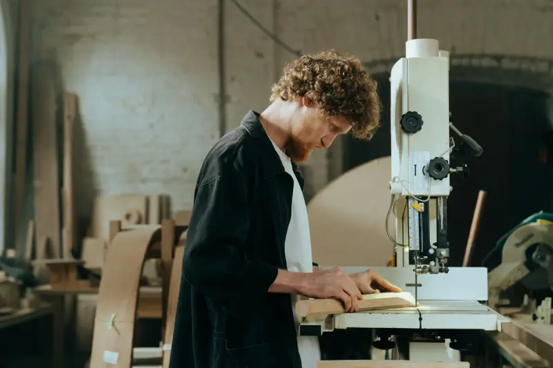Curly-haired craftsman guiding a wood piece through a band saw in a workshop