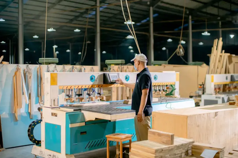 Worker setting up a multi-spindle boring machine beside stacks of wood parts