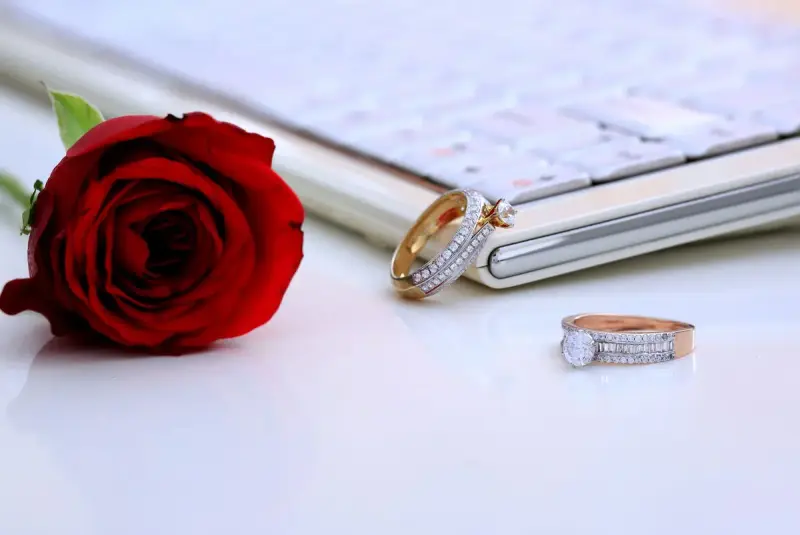 Two diamond rings on a white surface beside a red rose