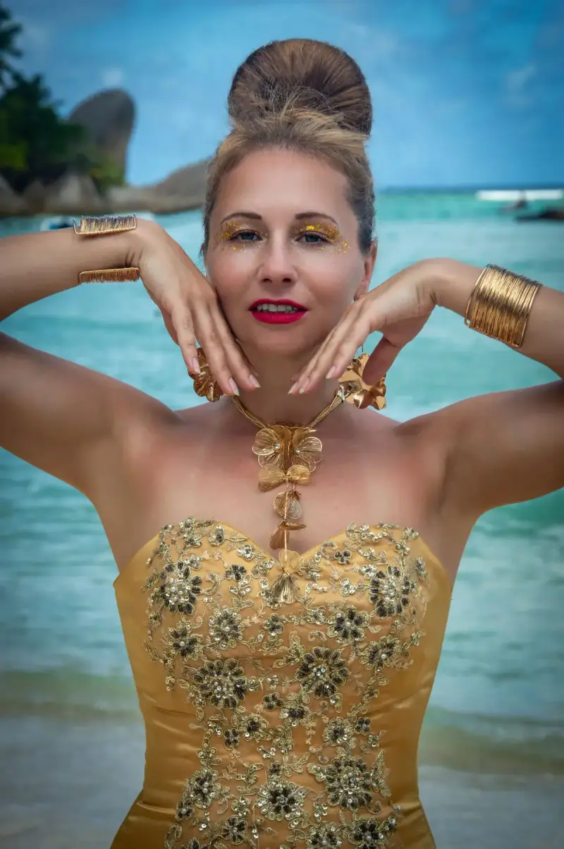 Woman in a gold embroidered dress wearing gold cuffs and a floral necklace by a turquoise sea