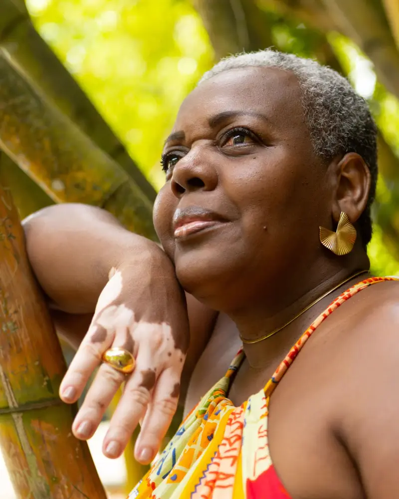 Older woman with gold fan earrings and a gold dome ring leaning on bamboo in sunlight