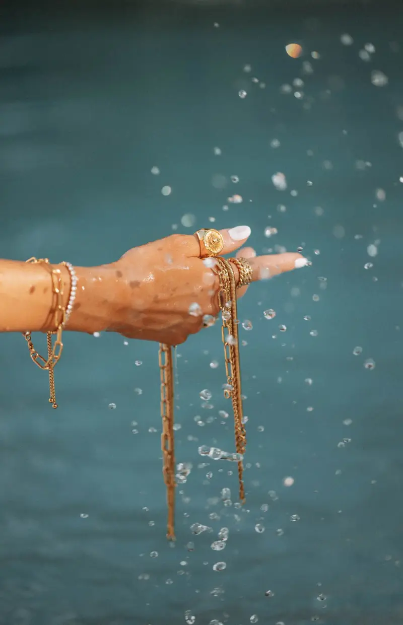 Hand wearing gold chain bracelets and rings over splashing turquoise water