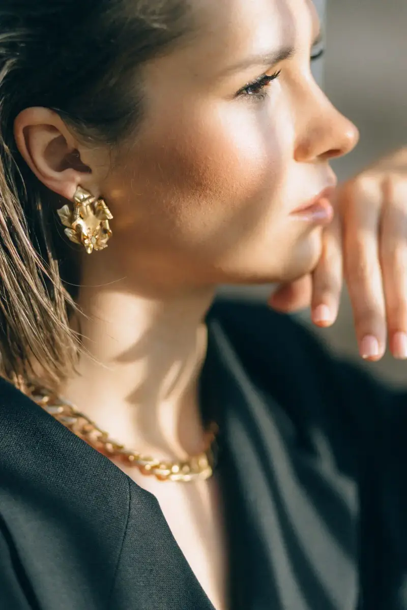 Woman in a black blazer with a sculpted gold earring and chunky gold chain in sunlight