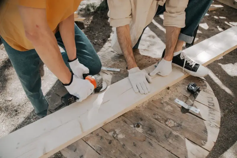 Two carpenters working a wooden plank with power tools outdoors