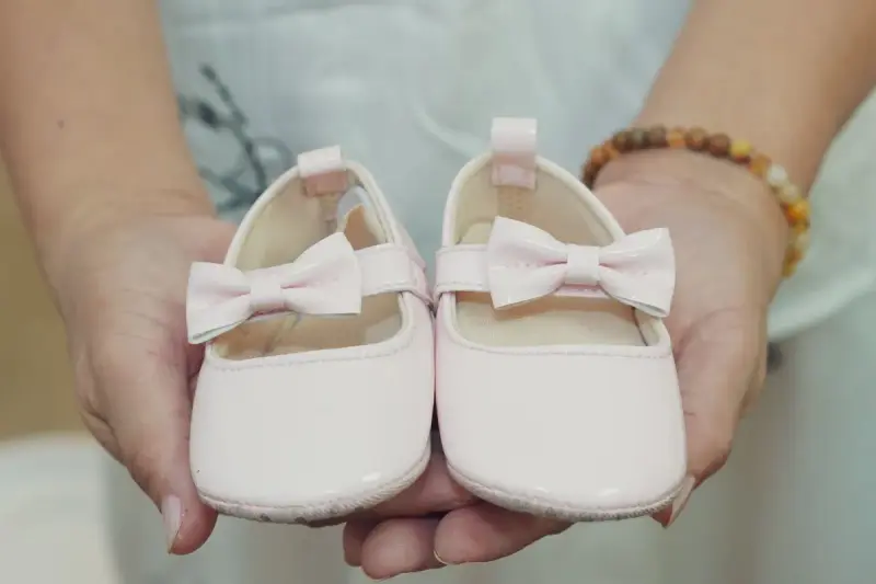 Hands holding a pair of pale pink baby shoes with bows