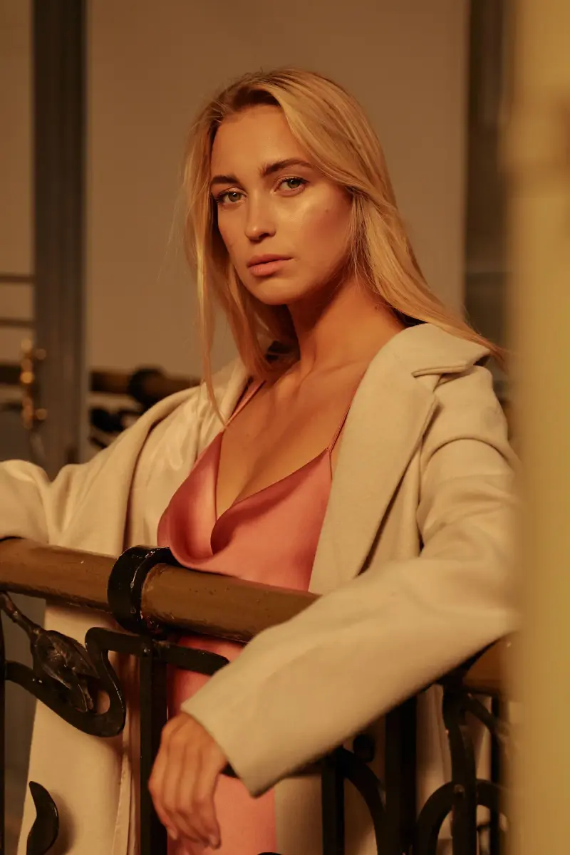 Woman in a camel coat and pink silk top leaning on a balcony rail