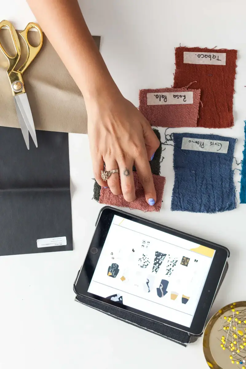 Hand choosing woven fabric swatches beside scissors and a tablet