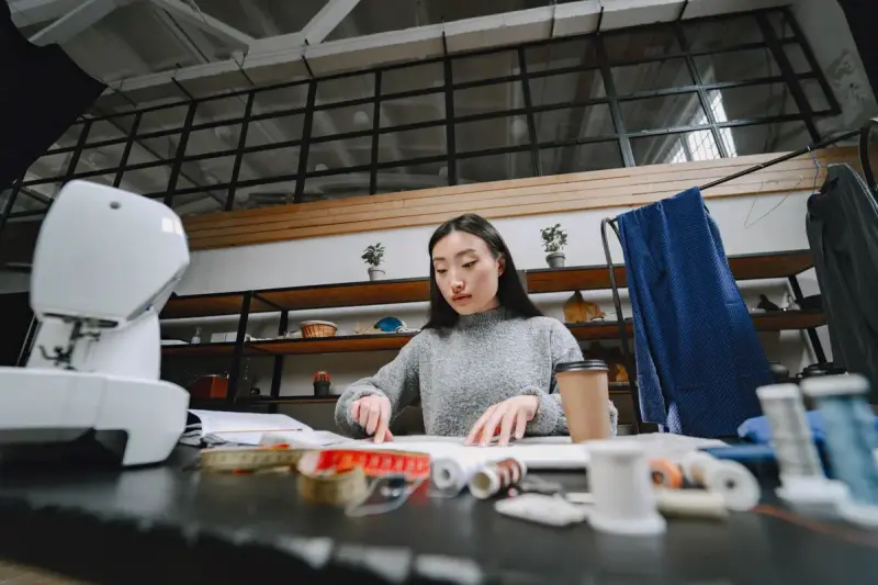 A fashion designer works at a desk surrounded by sewing tools and cloth