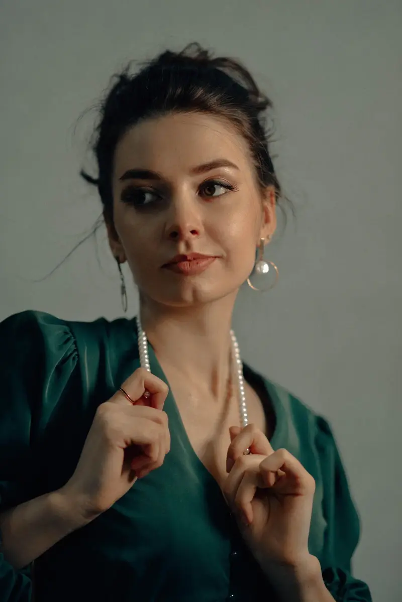 Woman in a green wrap dress holding a long pearl strand, wearing pearl drop earrings