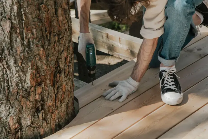 Worker in gloves drives screws into new deck boards beside a tree trunk