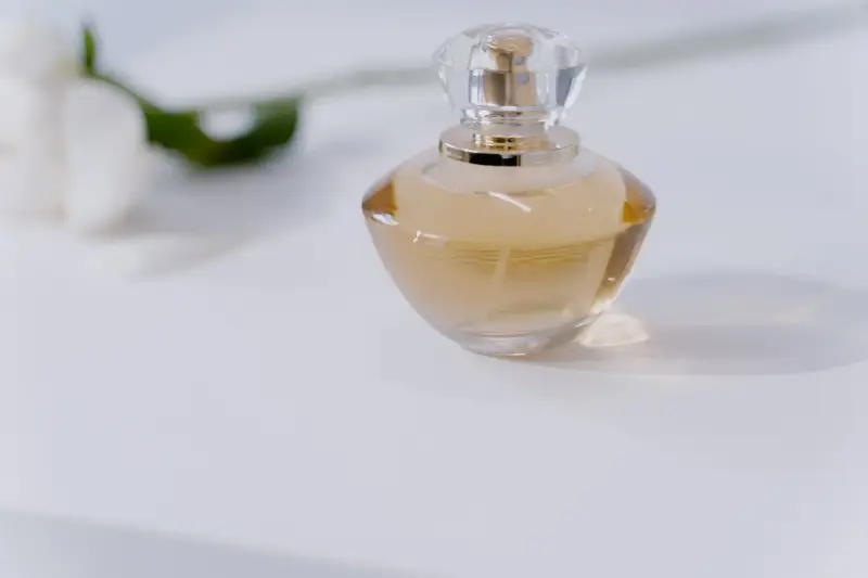 Round glass perfume bottle with a faceted cap on a white table beside a flower