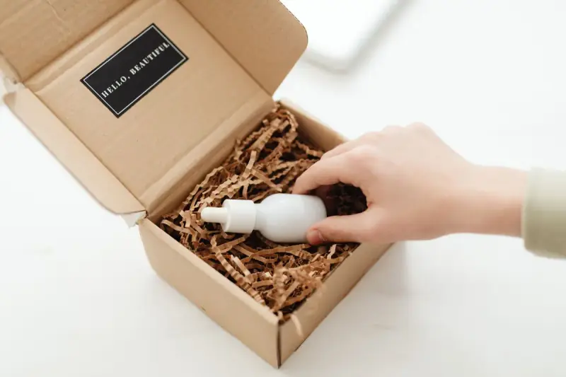 Hand lifting a white bottle from a kraft mailer box filled with paper shred