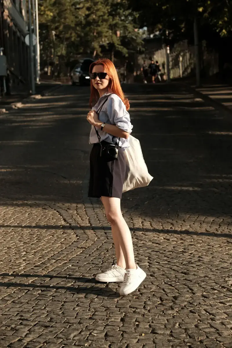 Red-haired woman in sunglasses, mini skirt and white sneakers on a sunlit cobblestone street