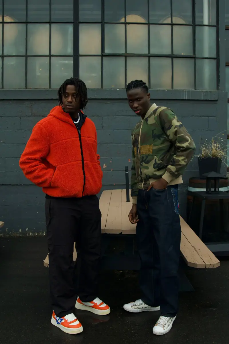 Two young men in streetwear, an orange puffer and a camo jacket, with sneakers