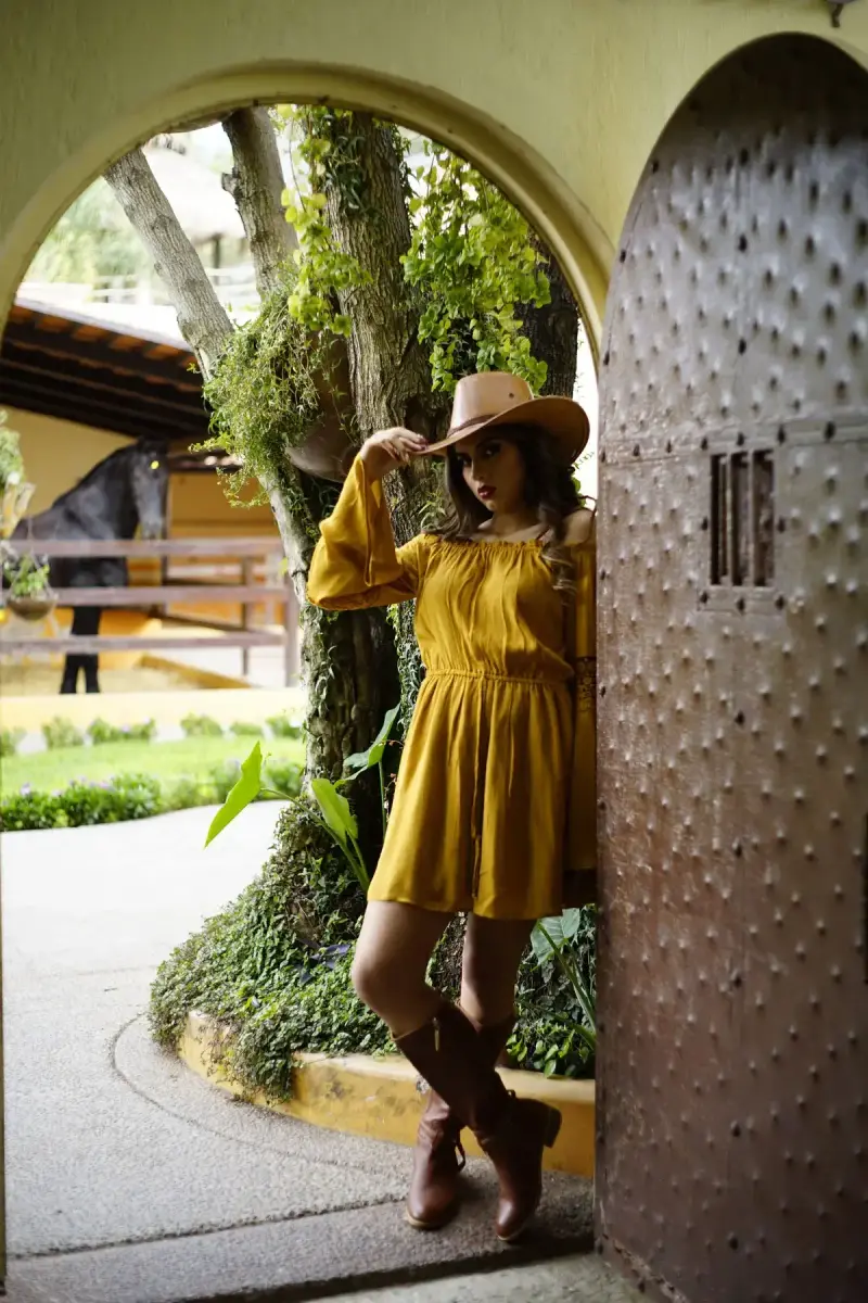 Woman in a mustard dress, cowboy hat and brown boots posing in an arched garden doorway
