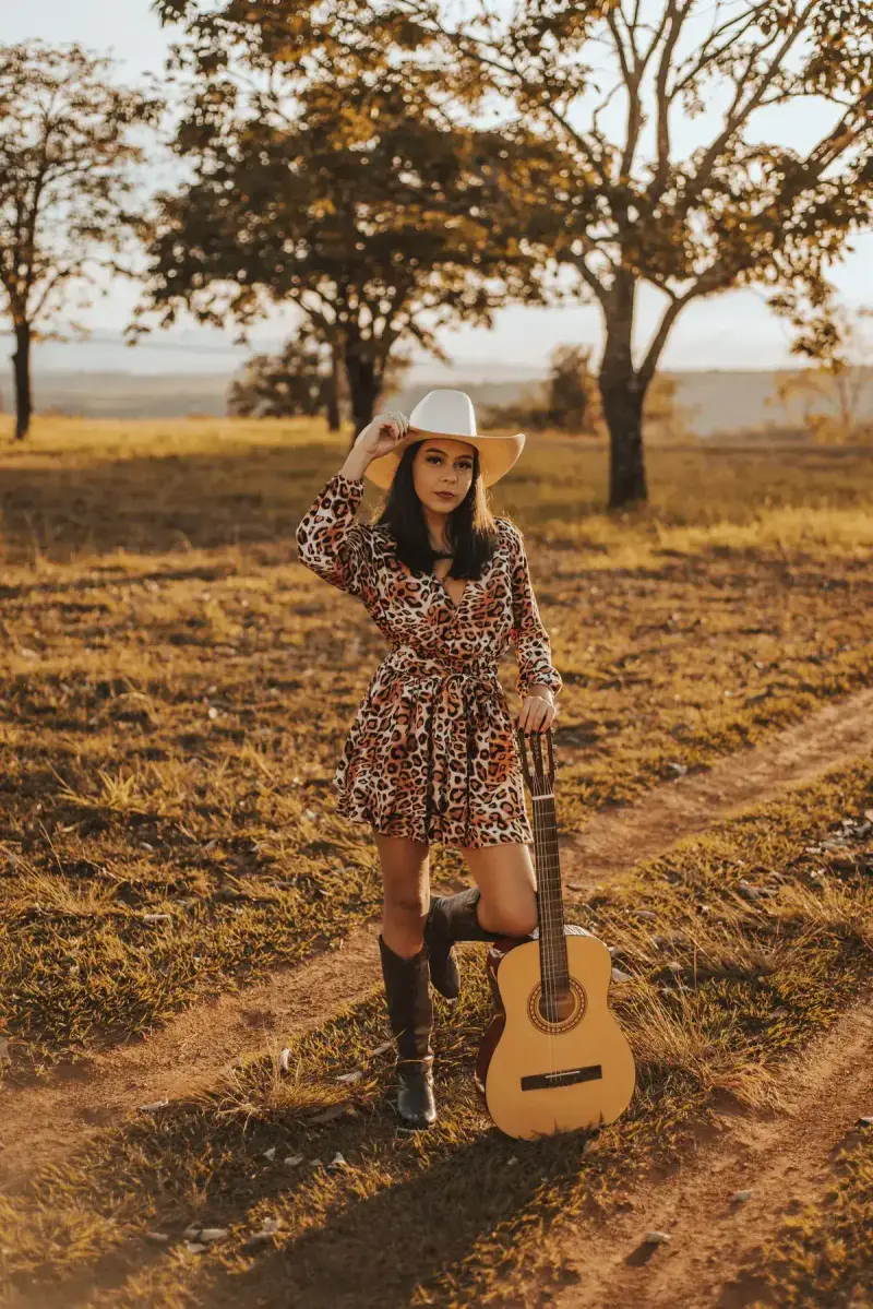 Woman in a printed mini dress, cowboy hat and boots holding an acoustic guitar at golden hour