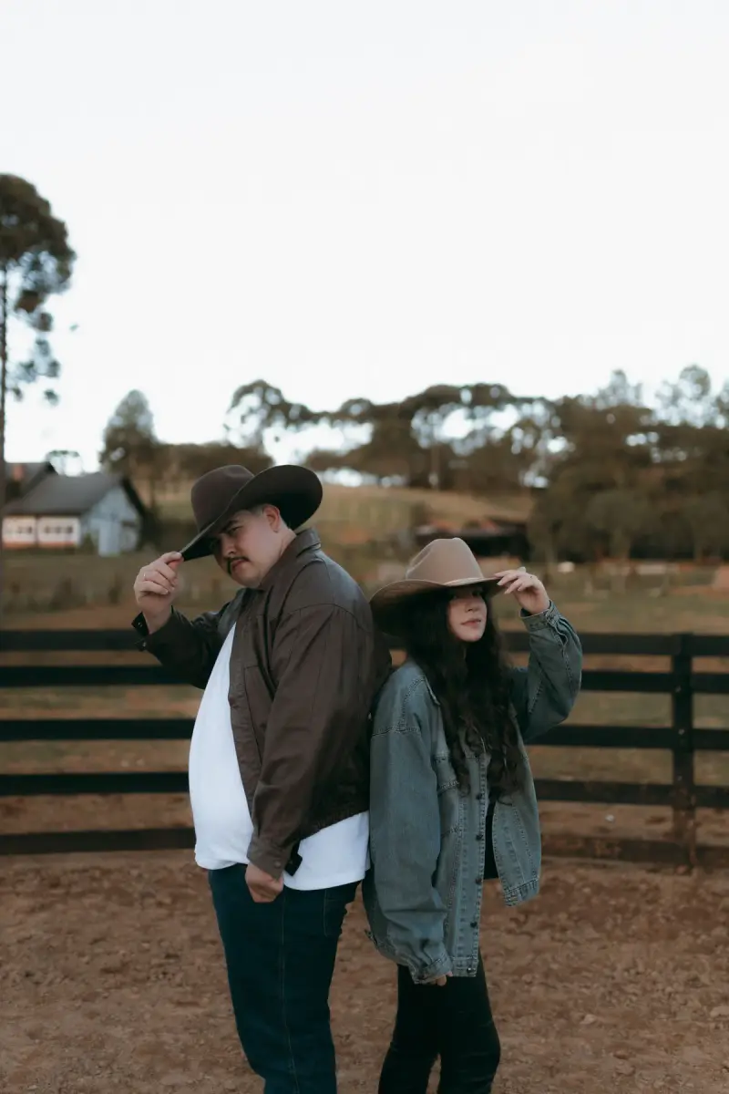 Couple in cowboy hats, a denim jacket and leather coat posing by a ranch fence