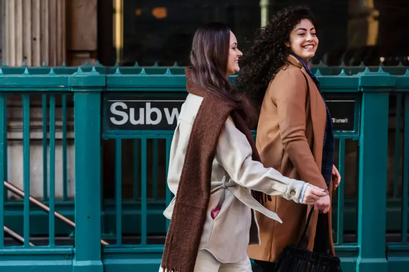 Two smiling women in camel and cream coats walking past a New York subway entrance