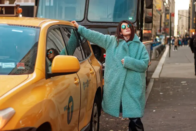 Woman in a mint teddy coat and sunglasses posing beside a yellow New York taxi