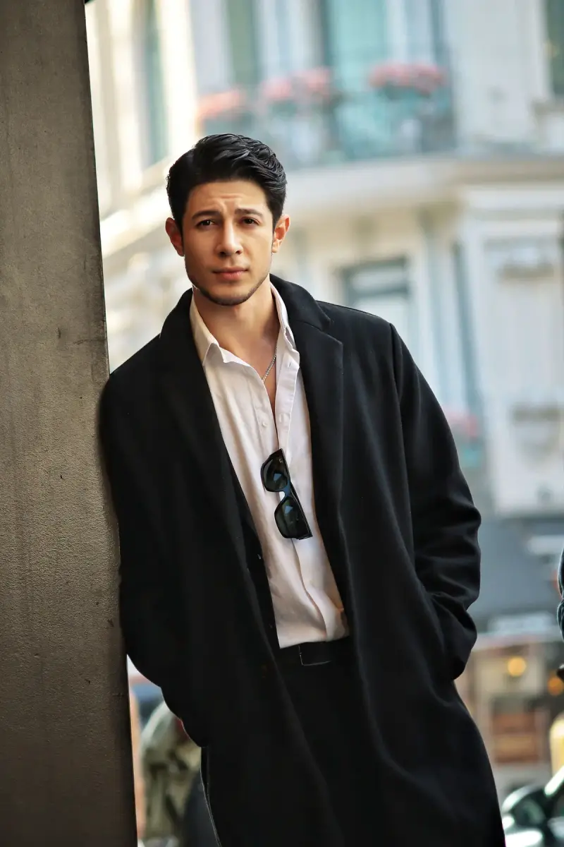 Young man in a long black overcoat and open white shirt leaning on a city building