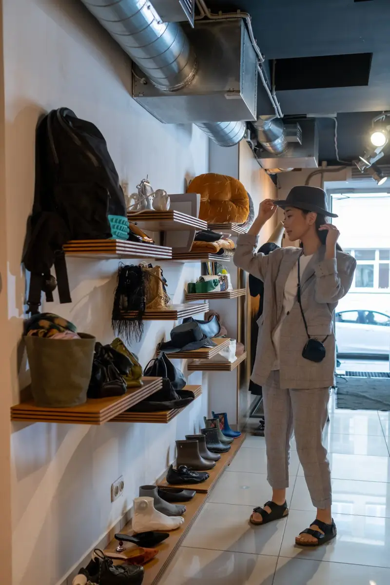 Woman trying on a hat in a boutique with bags and shoes on wall shelves