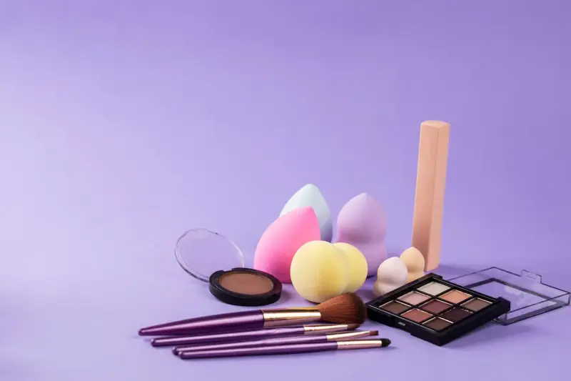Makeup sponges, brushes, a compact and an eyeshadow palette on a lilac backdrop