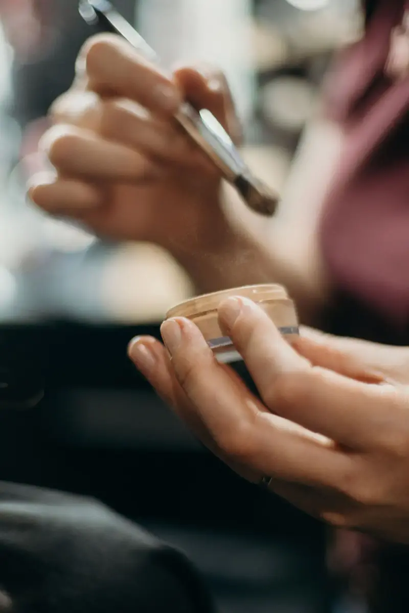 Hands holding a makeup brush and an open pot of loose powder