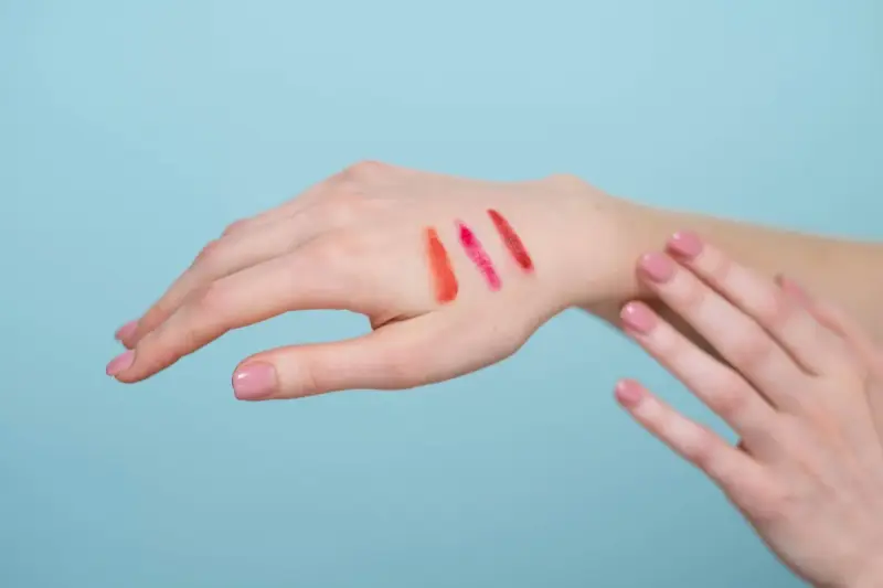 Red and pink lipstick swatches on the back of a hand against blue