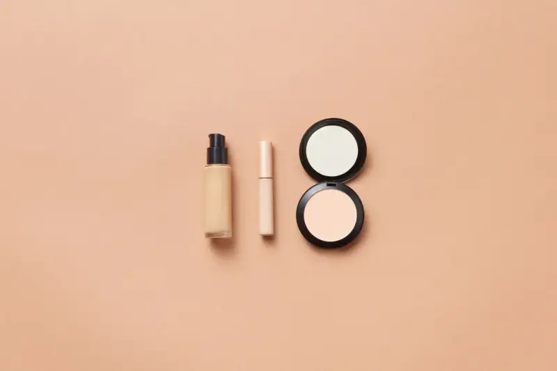 Foundation bottle, concealer and two compact powders on a peach background