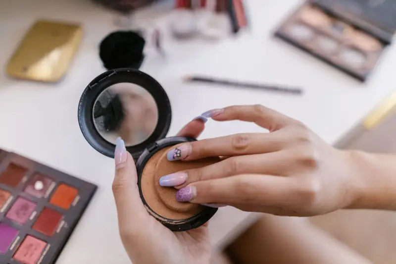 Hands opening a compact powder with a mirror above palettes on a white table