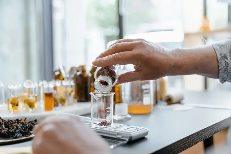 Hands adding dried botanicals to a glass beaker beside a scale and amber bottles