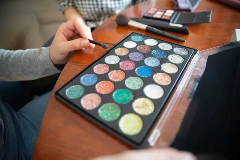 Hands holding a brush over a glitter eyeshadow palette on a wooden table