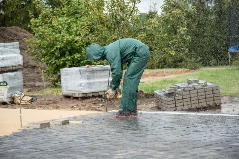 Worker in green rain gear laying gray paving stones beside stacked pallets of pavers