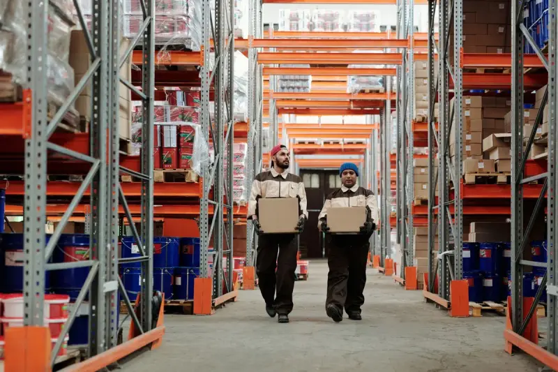 Two warehouse workers carrying cartons down an aisle between tall orange racks of stock